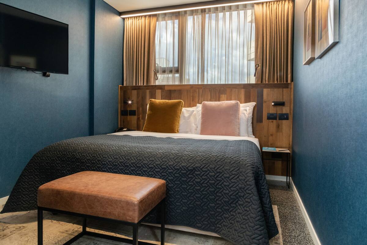 Habitación de hotel moderna con una cama grande, un banco, un televisor montado en la pared y un cabecero de madera.