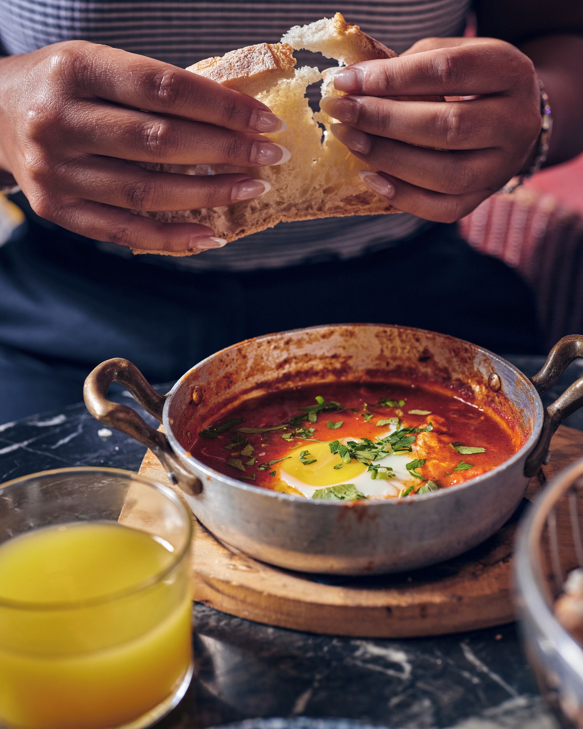 A person holding a piece of bread, a bowl of soup with an egg, and a glass of orange juice on a wooden board.