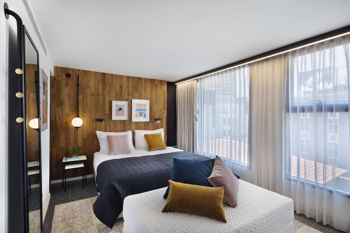 Habitación de hotel moderna con una cama grande, una mesita de noche pequeña, un espejo y vistas de la ciudad a través de cortinas translúcidas.