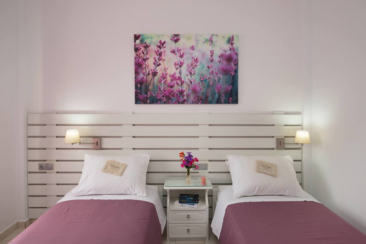 A hotel room with two beds, a nightstand with a lamp, a wall-mounted artwork, and a small table with a vase of flowers.