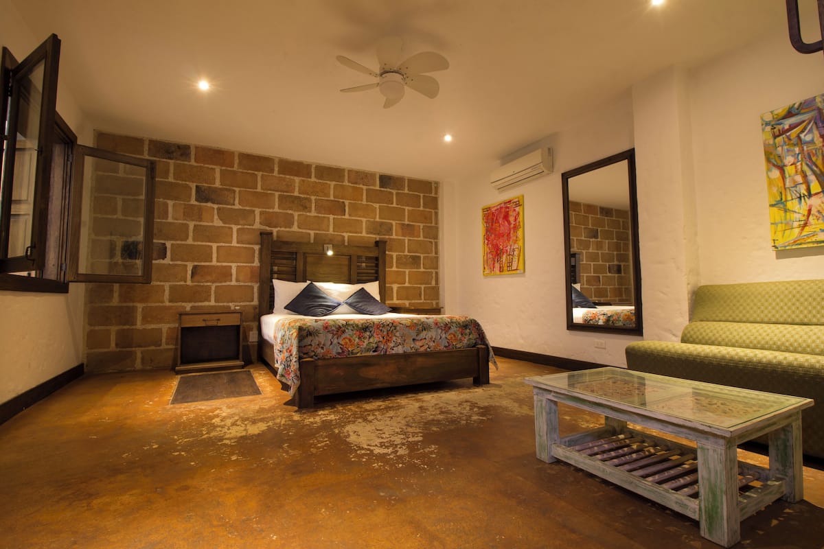A bedroom with a brick wall, a bed with a floral bedspread, a green sofa, a glass coffee table, and a ceiling fan.