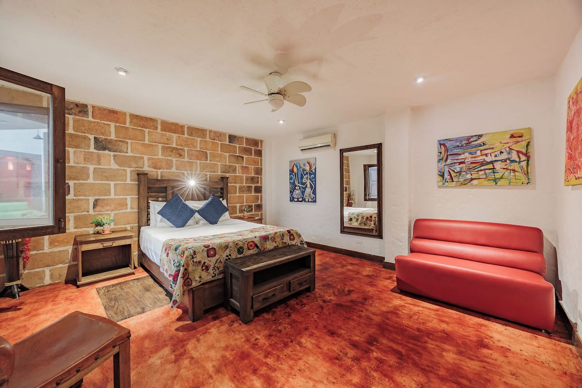 A bedroom with a brick wall, a ceiling fan, a bed with a colorful bedspread, a red sofa, and artwork on the walls.