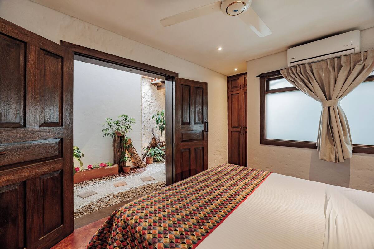A bedroom with a bed, a window with curtains, an air conditioning unit, and a door leading to an outdoor area with plants.