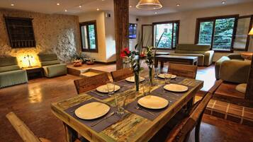 A dining area with a rustic wooden table set for four, surrounded by chairs. There is a sofa and a television in the background.