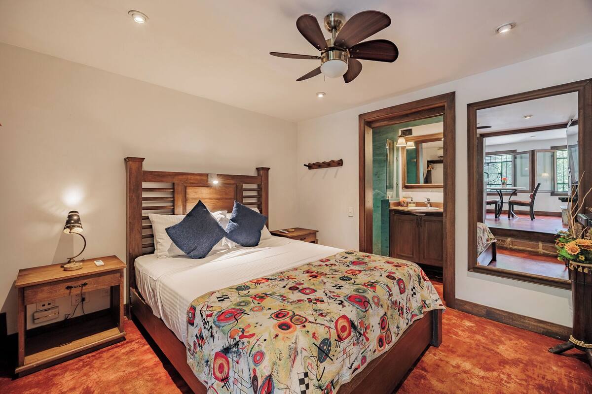A bedroom with a bed, wooden headboard, bedside table with lamp, ceiling fan, and large mirror.