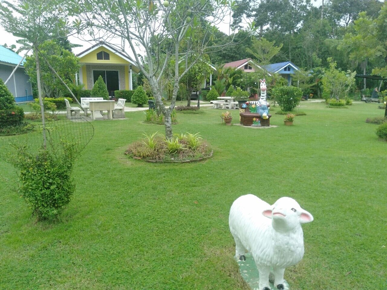 A grassy area with a white sheep statue, a small tree, and a house with a blue roof in the background.