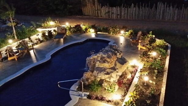 Vista nocturna de una piscina con una formación rocosa, rodeada de sillas y plantas, con una valla de madera al fondo.