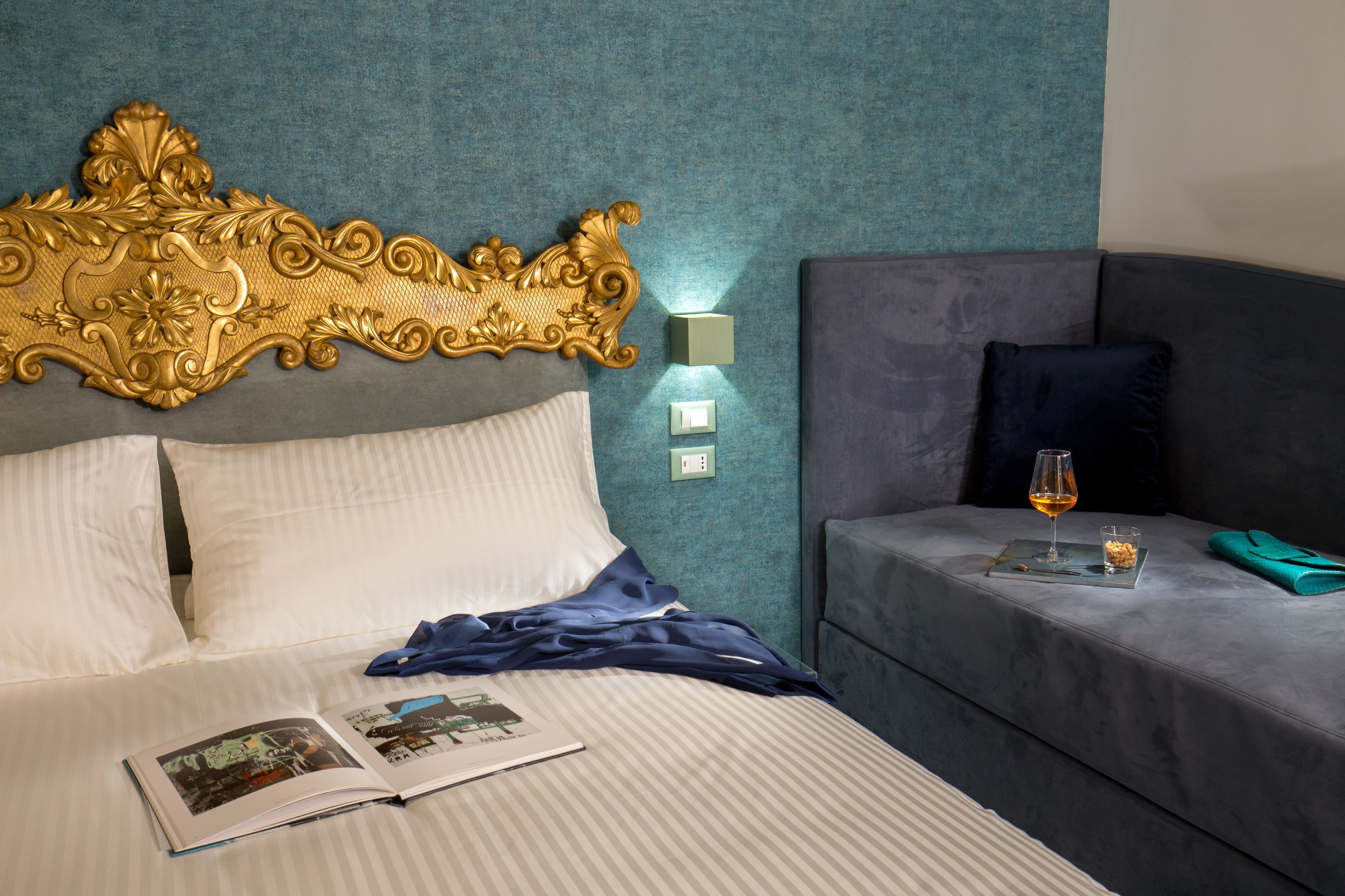 Une chambre d’hôtel avec une grande tête de lit décorée, un lit recouvert de draps blancs, une table de chevet avec un verre de vin et des en-cas, et un canapé gris.