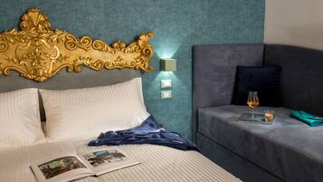 A hotel room with a large ornate headboard, a bed with white linens, a bedside table with a glass of wine and snacks, and a grey sofa.