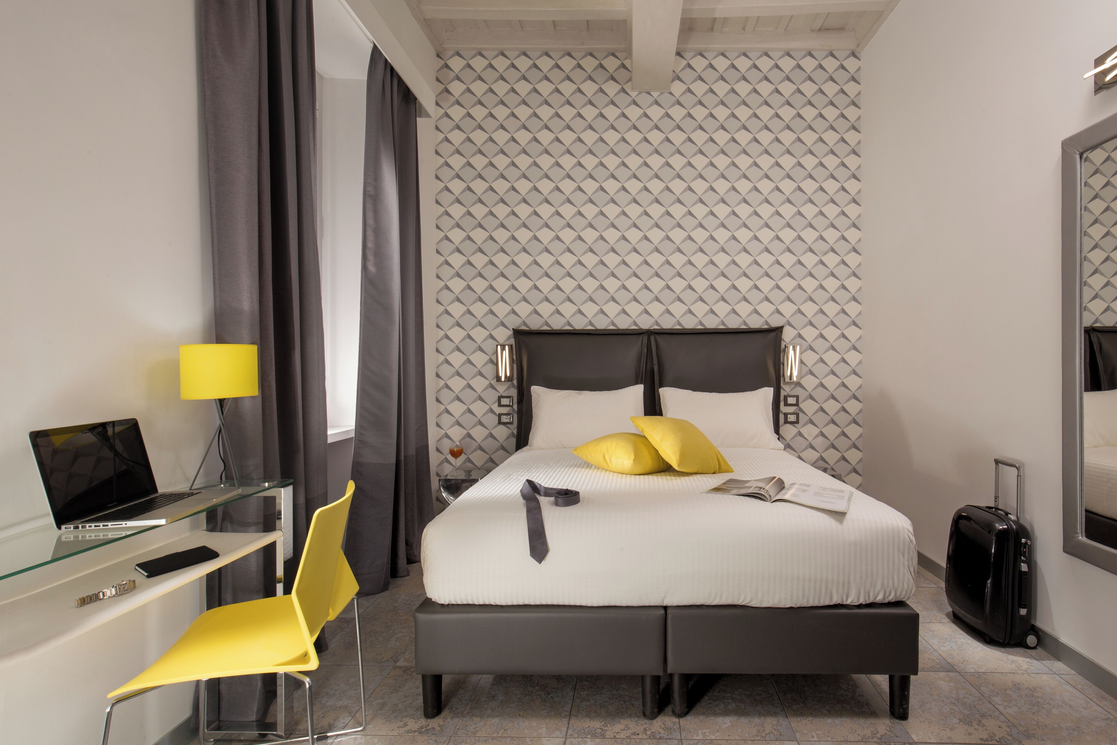 A modern hotel room with a bed, a desk with a laptop, a chair, a suitcase, and a mirror.