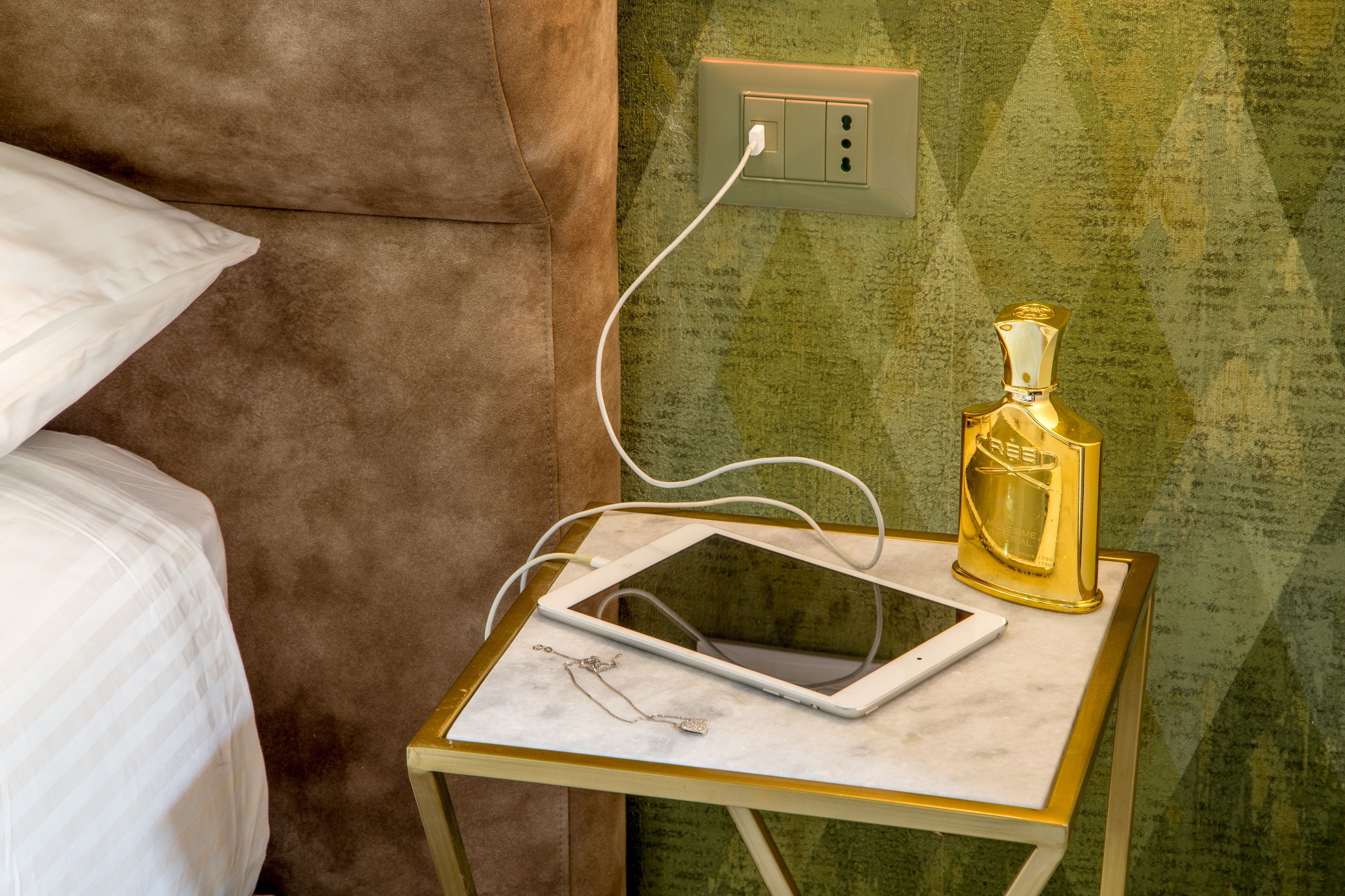 A neatly made bed with a white pillow, a small bedside table with a tablet and a gold bottle, and a green textured wall.