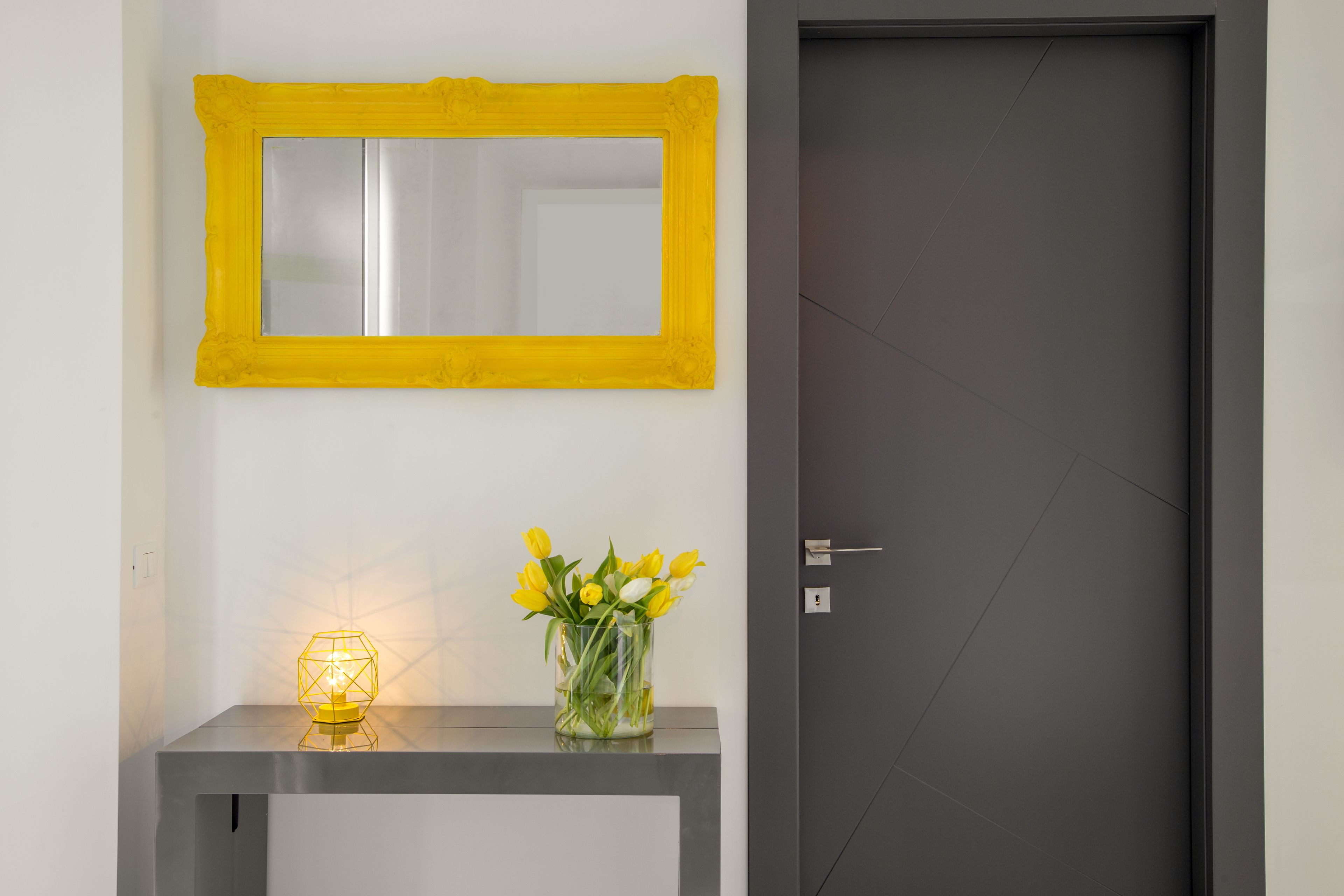 Een kamer met een spiegel, een vaas met gele bloemen en een tafel met een geometrisch lampje.