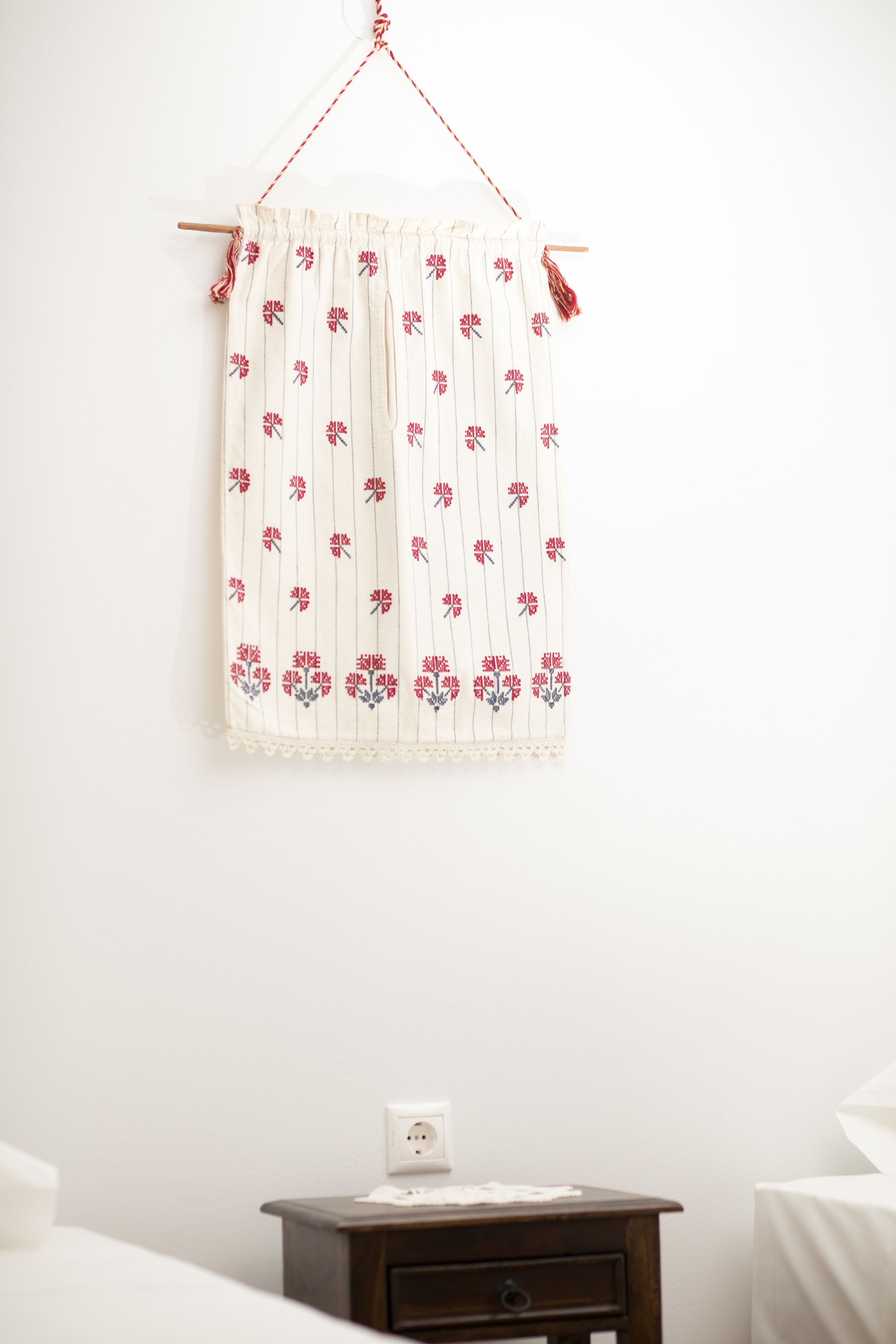 A white wall with a hanging curtain featuring a floral pattern. Below the curtain, there is a wooden nightstand with a white lamp and a power outlet.