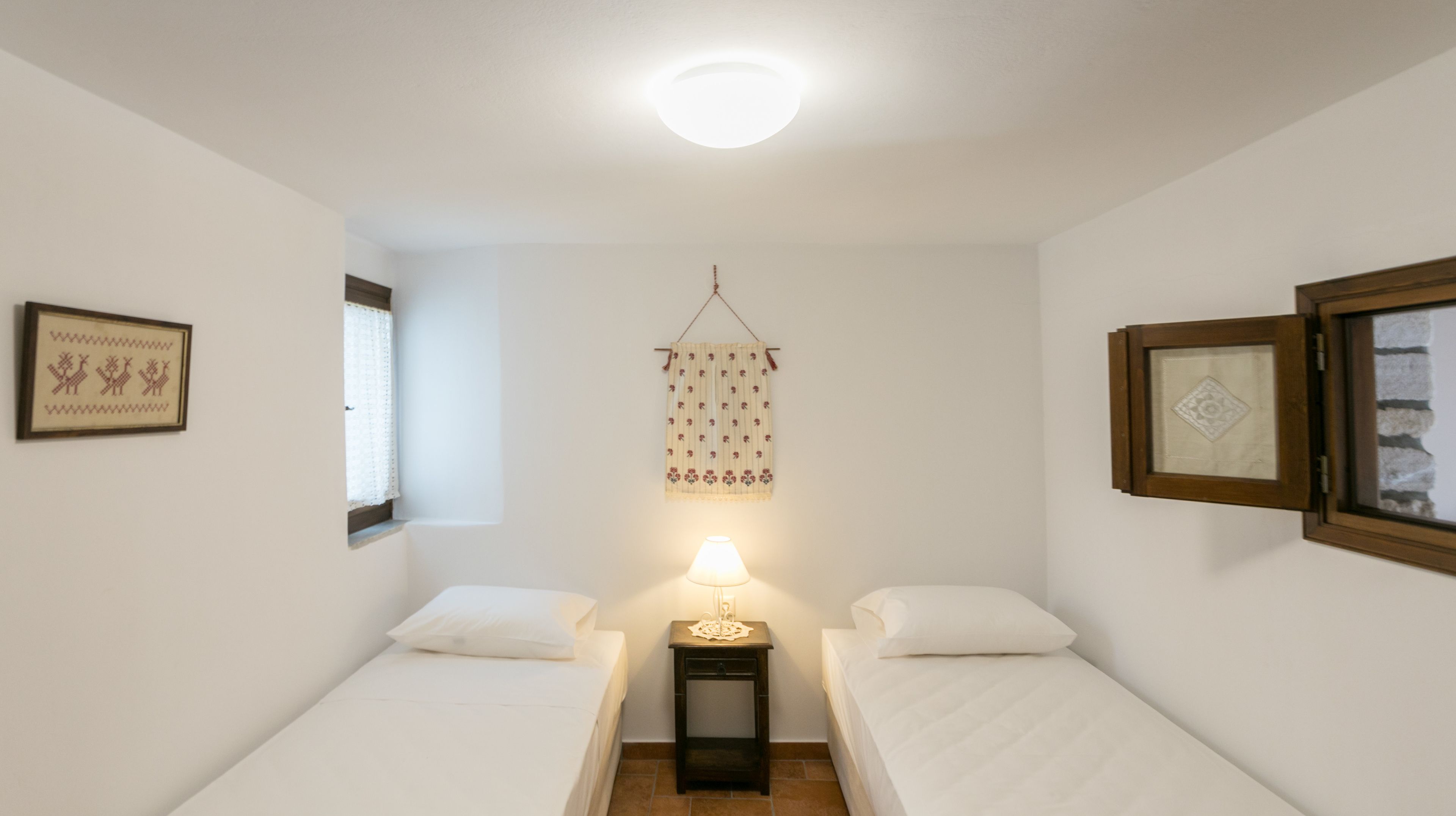 Una stanza piccola e pulita con due letti singoli, un comodino con una lampada e un decorativo tessile appeso.