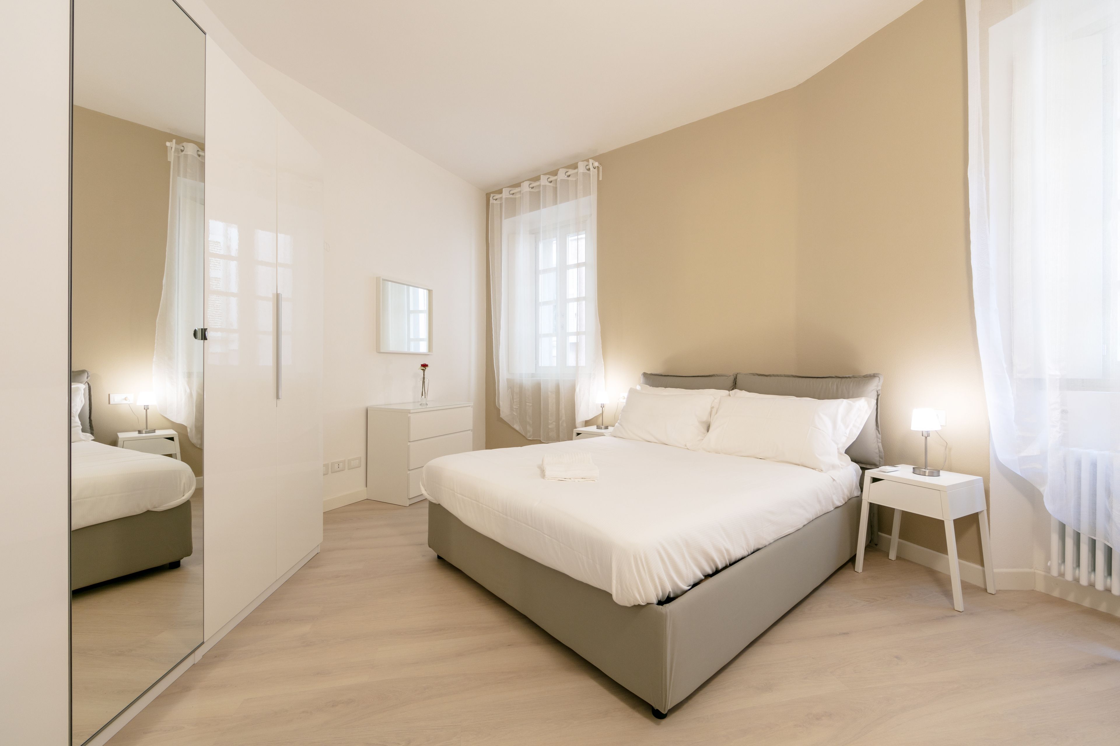 A modern bedroom with a large bed, bedside tables, a mirror, and a window with sheer curtains.