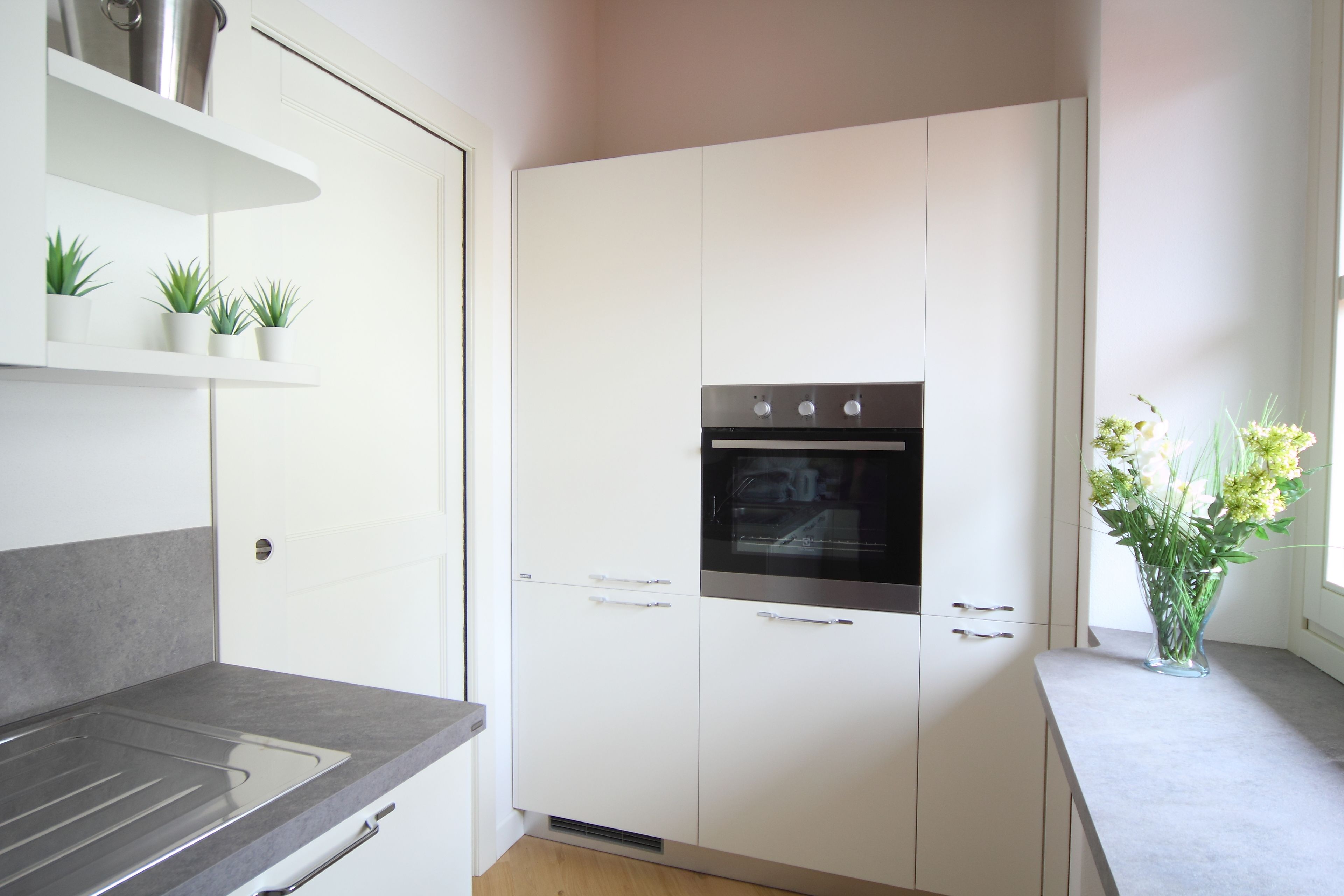 A modern kitchen with white cabinets, a built-in oven, and a countertop with a sink. There are small potted plants on a shelf above the cabinets.