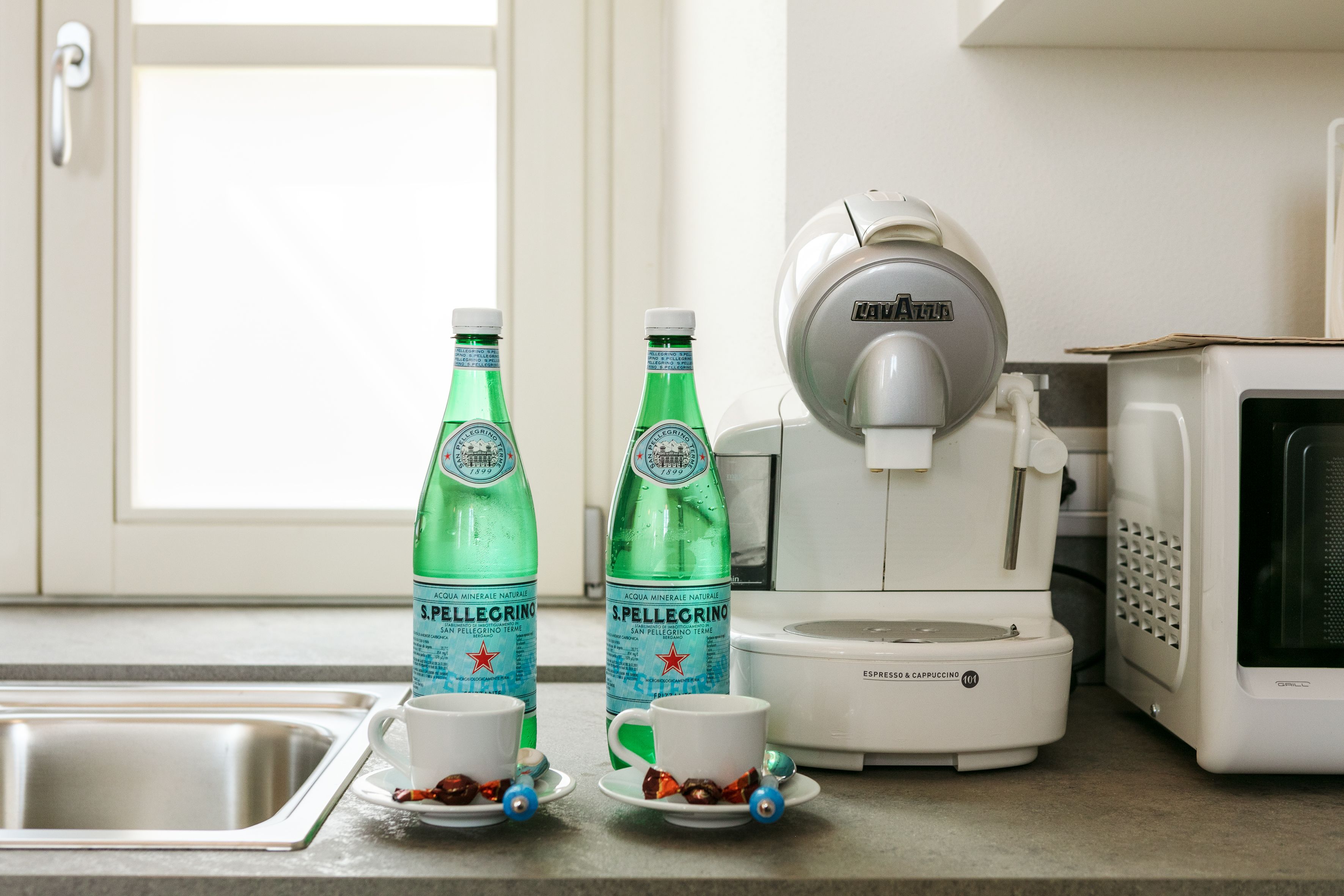Een keuken met een koffieapparaat, twee flessen Pellegrino water en twee kopjes op het aanrecht.