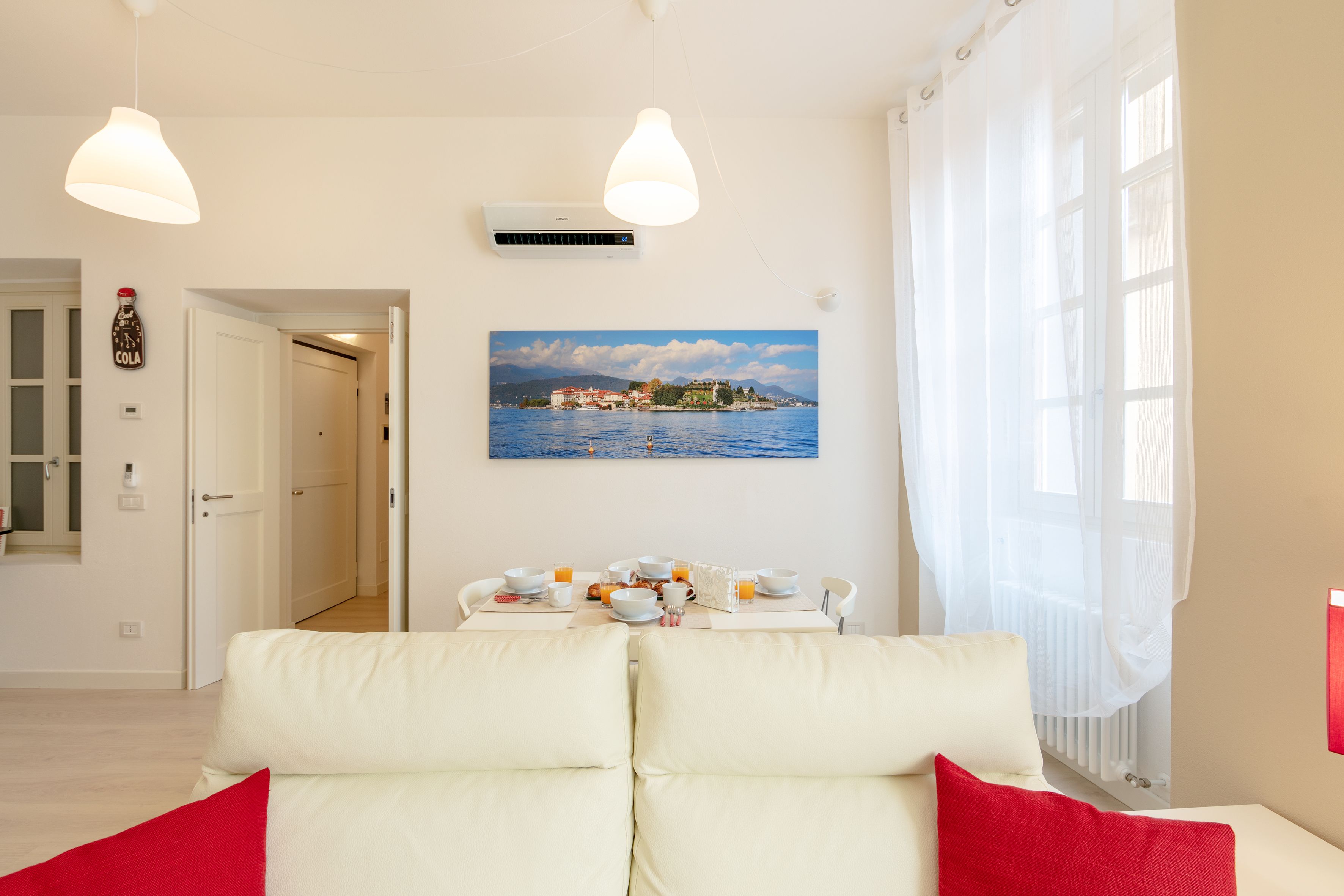 A modern living room with a white sofa, a dining table set with breakfast items, and a painting of a coastal scene.