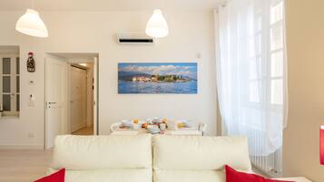 A modern living room with a white sofa, a dining table set with breakfast items, and a painting of a coastal scene.