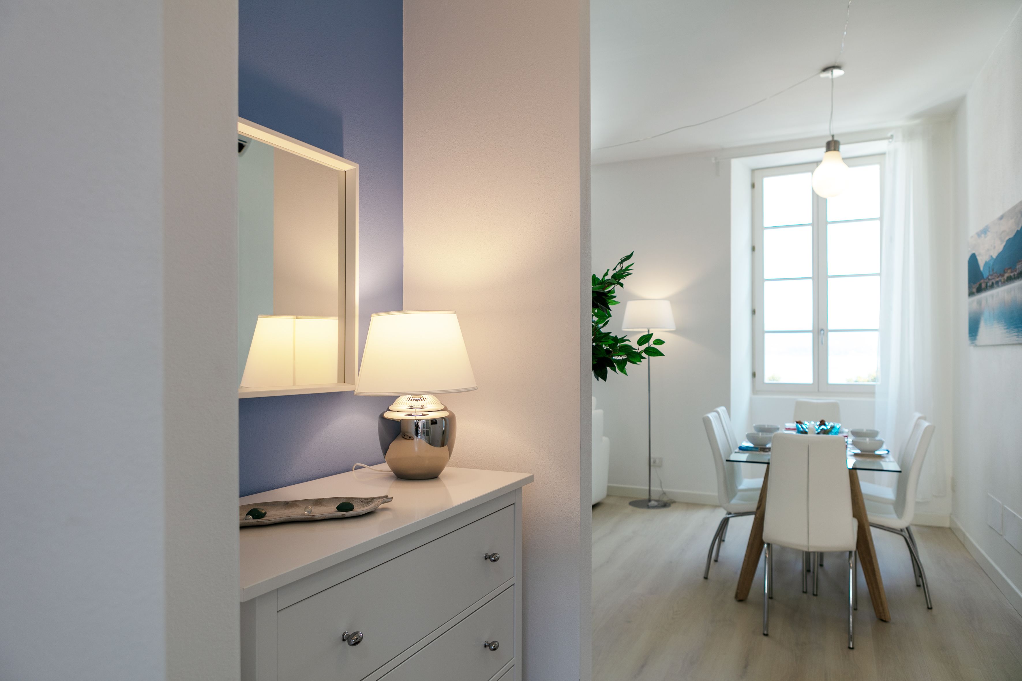 A modern room with a white dresser, two lamps, a dining table with chairs, and a large window with natural light.