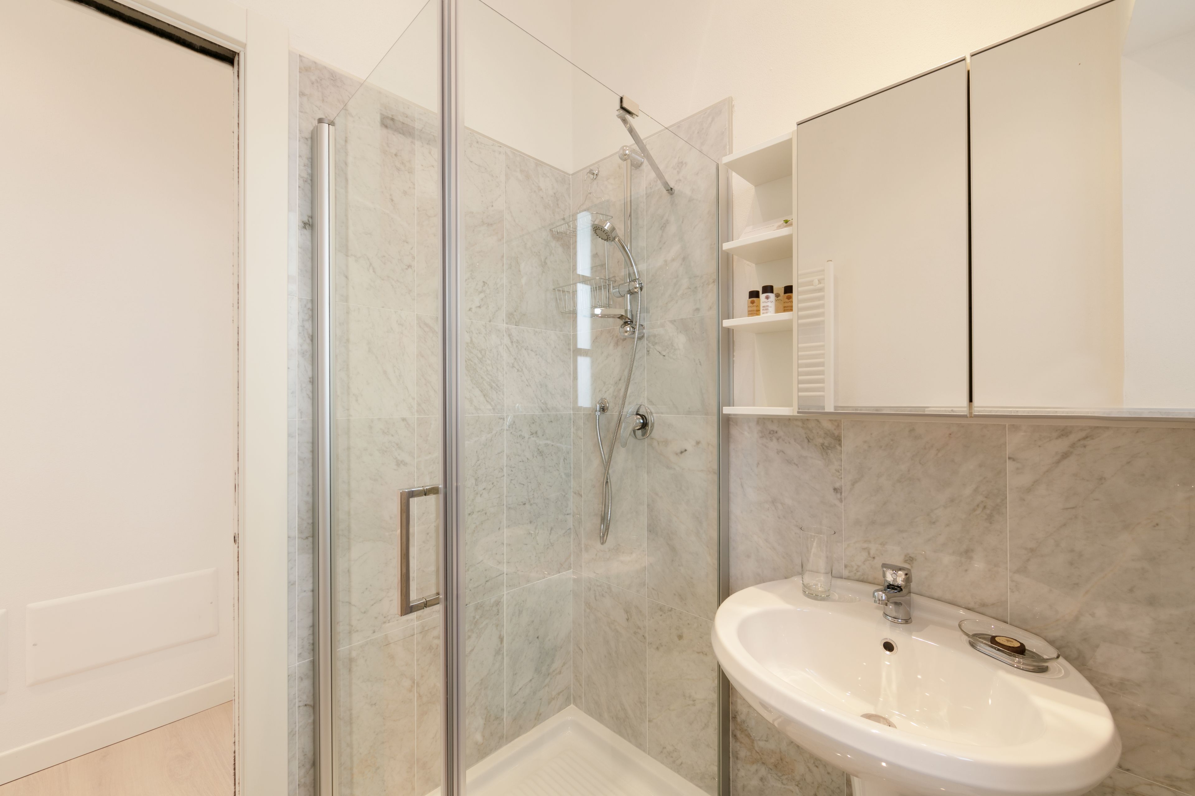 Un baño moderno con ducha de vidrio, lavamanos blanco y pared con azulejos de mármol.