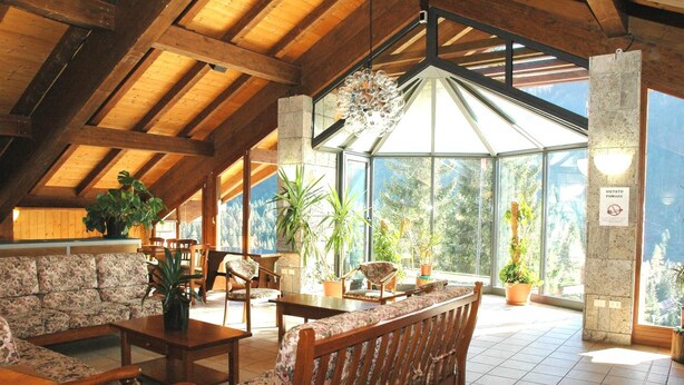A spacious indoor area with wooden beams, a high ceiling, and large windows offering a view of trees. There are multiple seating areas with floral upholstery and potted plants.