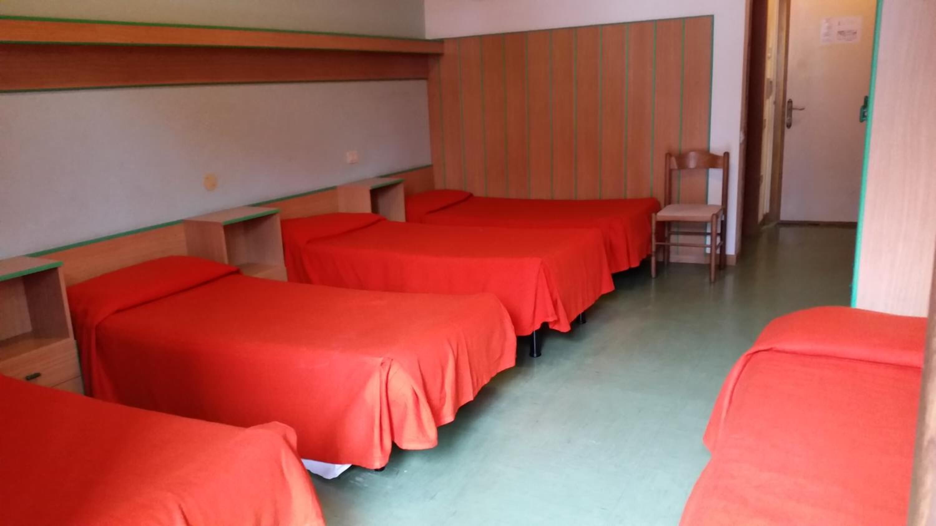 A dormitory room with four beds, each covered with red sheets, wooden headboards, and a green floor.