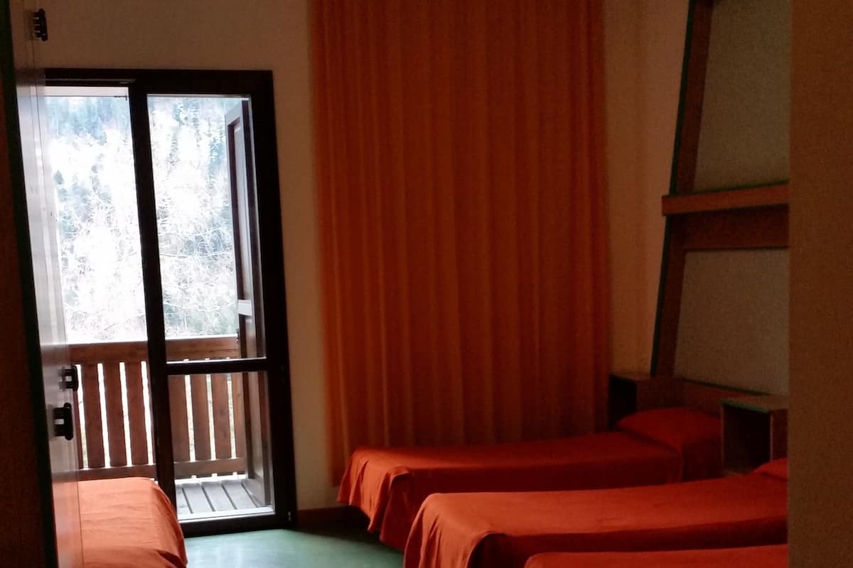 A room with two beds, a wooden bench, a large window with curtains, and a view of trees outside.