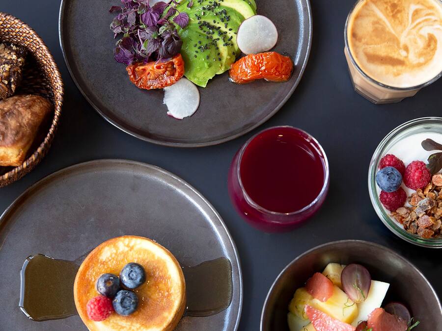 Una colazione con diversi piatti, tra cui pancake, avocado, frutti di bosco e un latte macchiato.