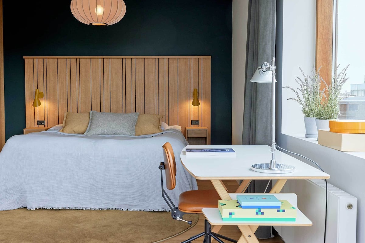 A modern bedroom with a wooden headboard, a desk with a chair, a bedside table, and a window with a view of the city.