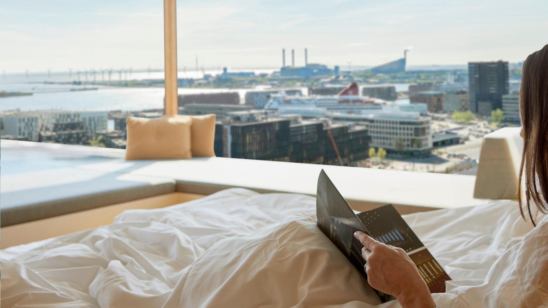 Una persona acostada en una cama en una habitación con un ventanal grande que da a una vista panorámica de la ciudad.