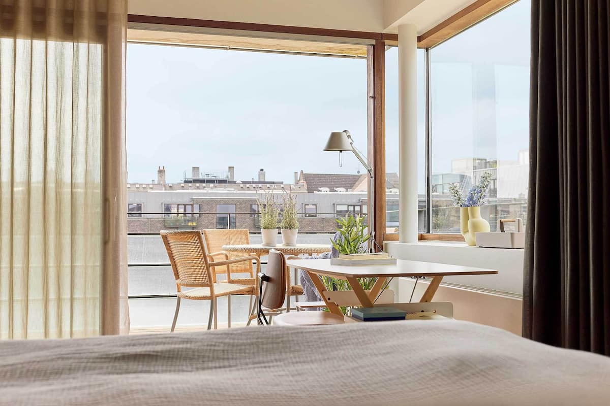 A modern hotel room with a large window, a dining table with chairs, and a view of the cityscape.
