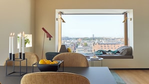 A modern living room with a large window, a dining table with candles, a bowl of lemons, and a view of a cityscape. - Charlottehaven (Copenhagen)