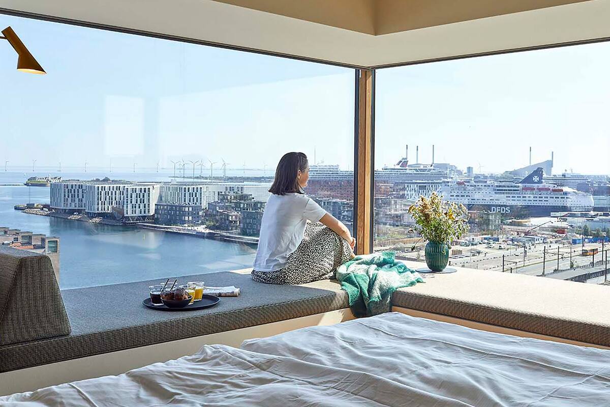 A modern hotel room with a large window overlooking a cityscape, including a harbor and buildings.