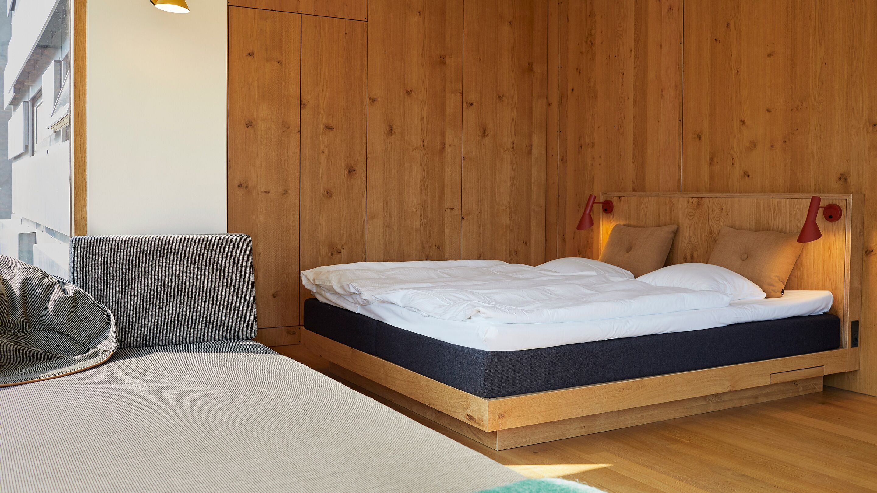A modern bedroom with a wooden headboard, a single bed with white bedding, a grey sofa, and a red wall lamp.