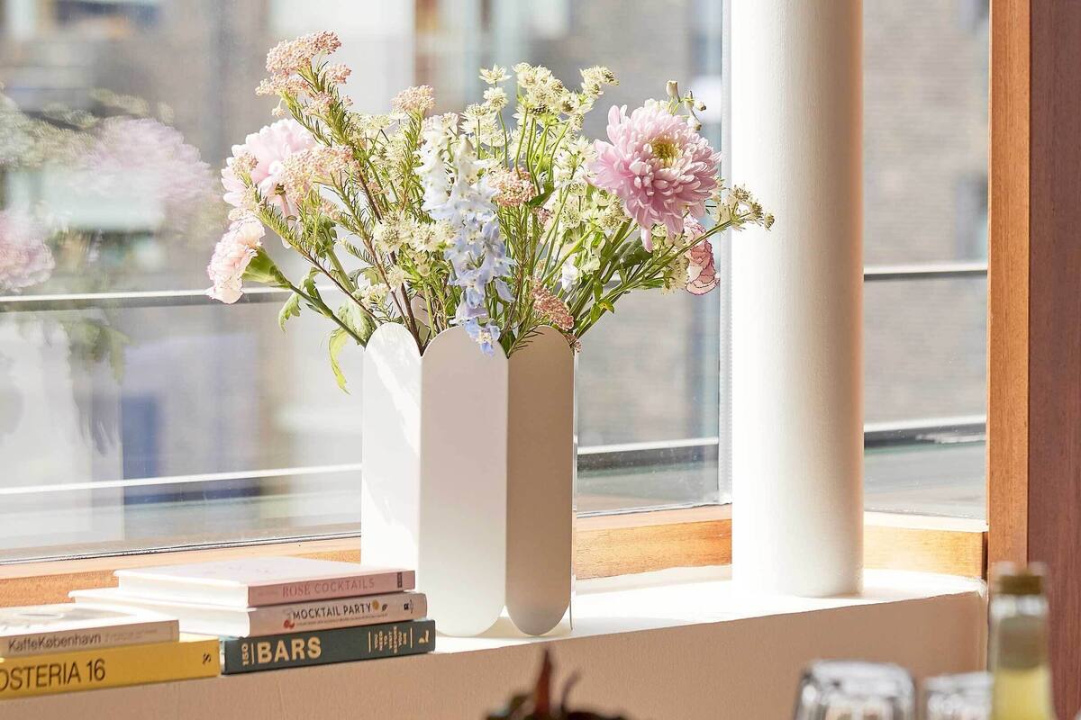 A white vase with flowers on a windowsill, next to a stack of books.