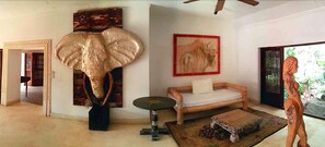 A room with a large elephant sculpture, a sofa, a small round table, and a wooden chair. - White Elephant Sea & Art Lodge (Malindi)