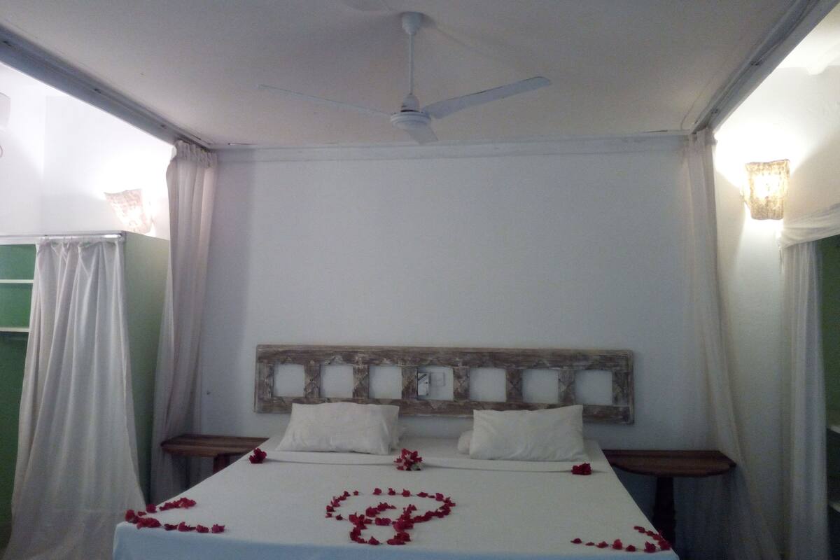 Un lit avec du linge de lit blanc et des pétales de rose disposés en forme de cœur, un ventilateur de plafond et une tête de lit en bois.