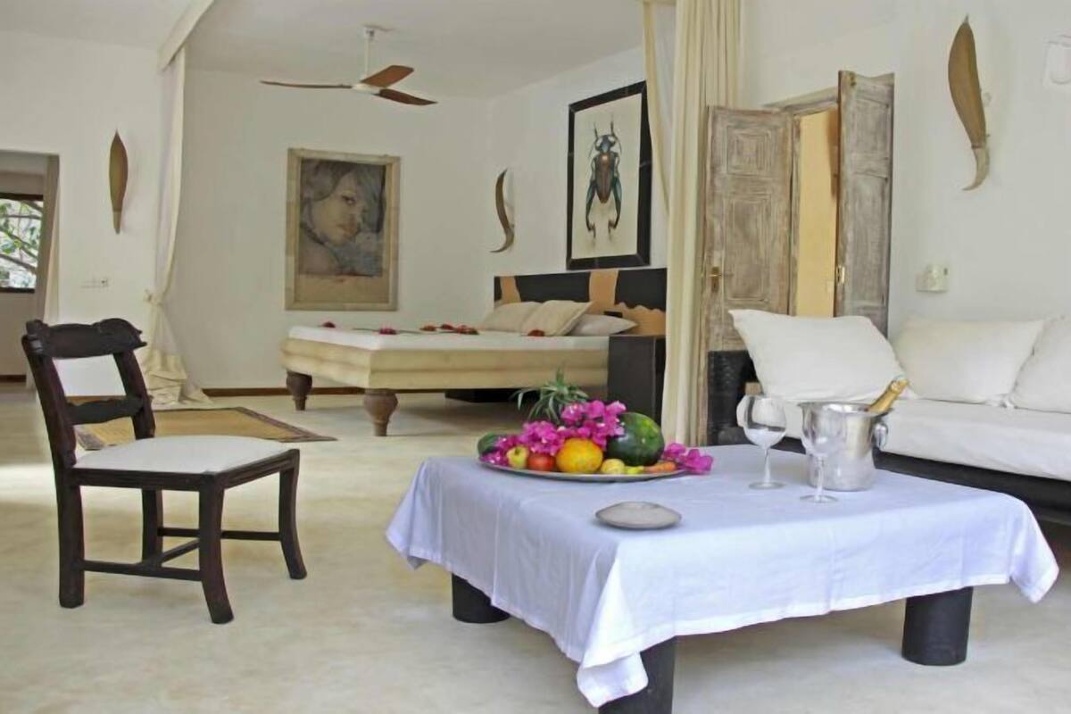 Une chambre à coucher avec un lit, un canapé blanc, une chaise en bois, une table avec un panier de fruits et un verre à vin.