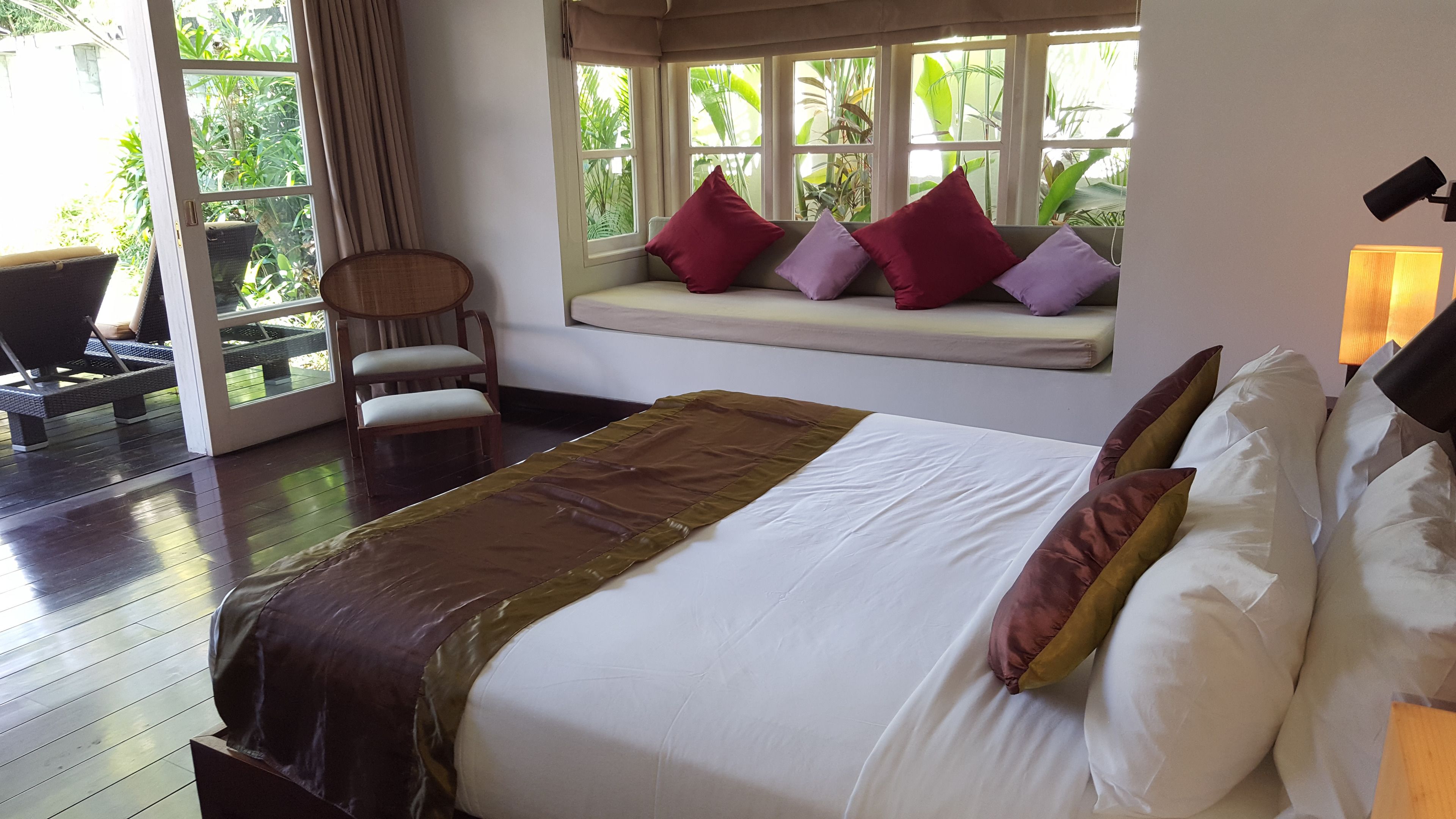 A bedroom with a large bed, a window seat with cushions, a chair, and a view of greenery outside.