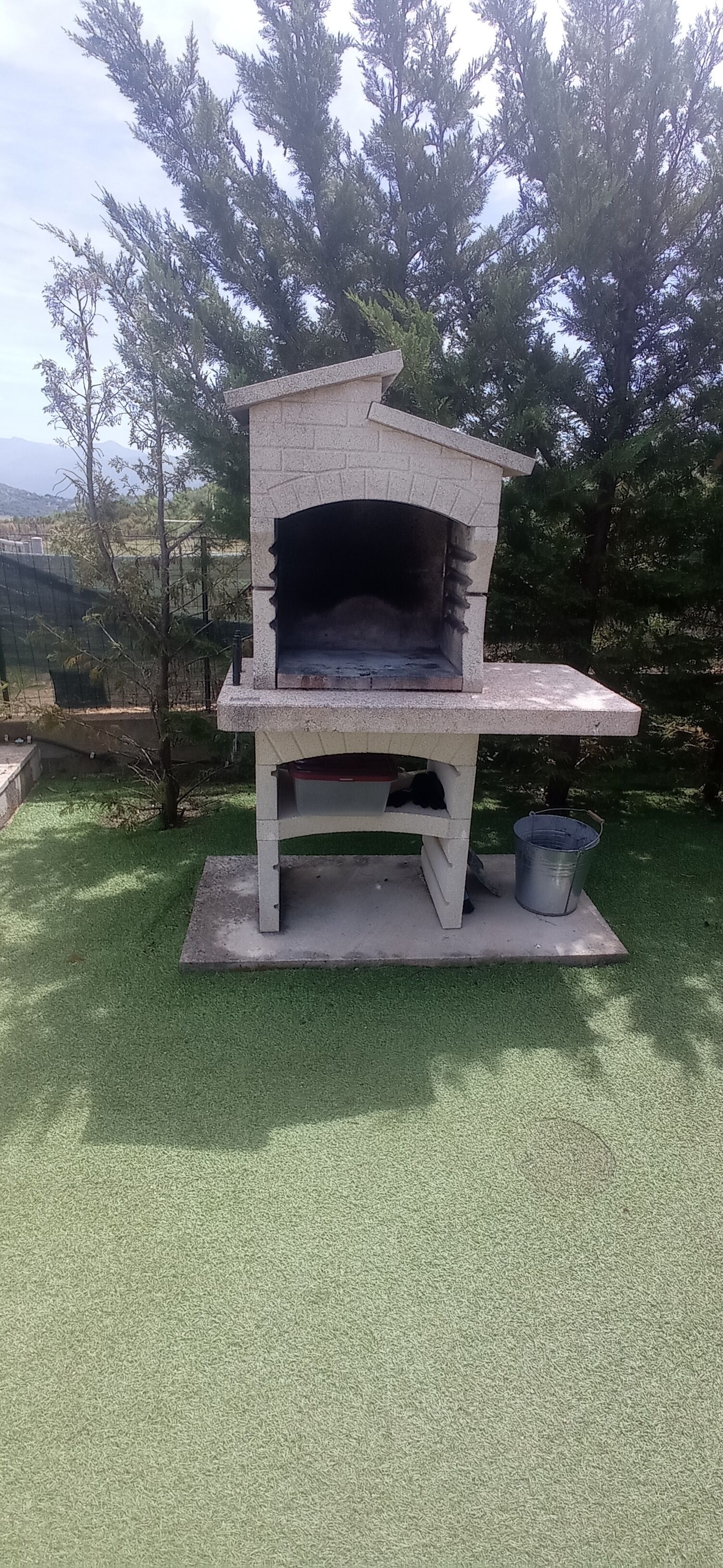 Fornet de pizza de pedra a l'aire lliure amb xemeneia, col·locat sobre una base de formigó, envoltat d'herba verda i arbres.