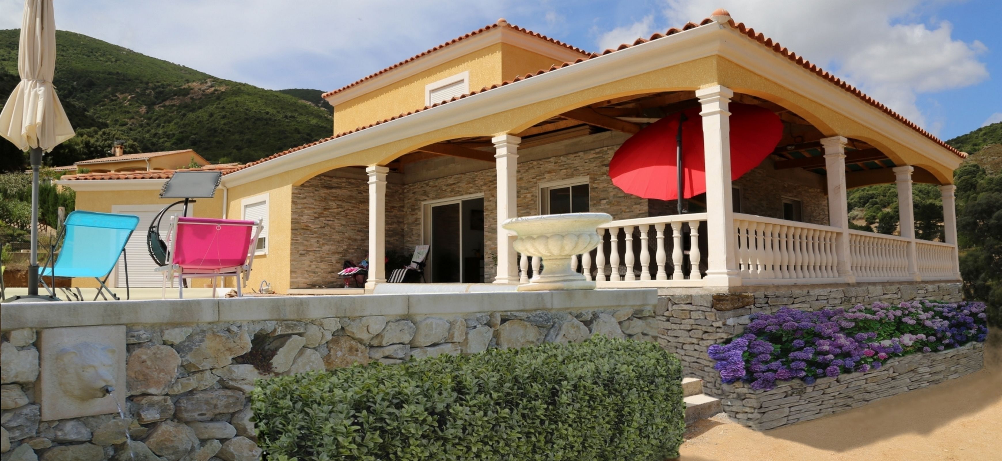 Una casa con fachada de piedra, un porche cubierto con un paraguas rojo y mobiliario para exteriores.
