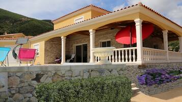 Una casa con fachada de piedra, un porche cubierto con un paraguas rojo y mobiliario para exteriores.