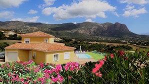 Una villa amarilla con piscina, rodeada de vegetación exuberante y montañas al fondo.