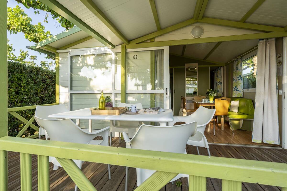Uma área externa coberta com mesa e cadeiras, deck de madeira e vista para o verde ao redor.