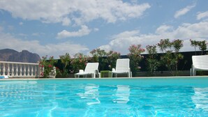 Una piscina azul cristalina con sillas de descanso blancas y flores rosadas al fondo.