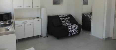 Ett kompakt kök med en mikrovågsugn, en liten arbetsbänk och ett skåp. Där finns en svart soffa med en pläd med en QR-kod, en vit papperskorg och en stor spegel.