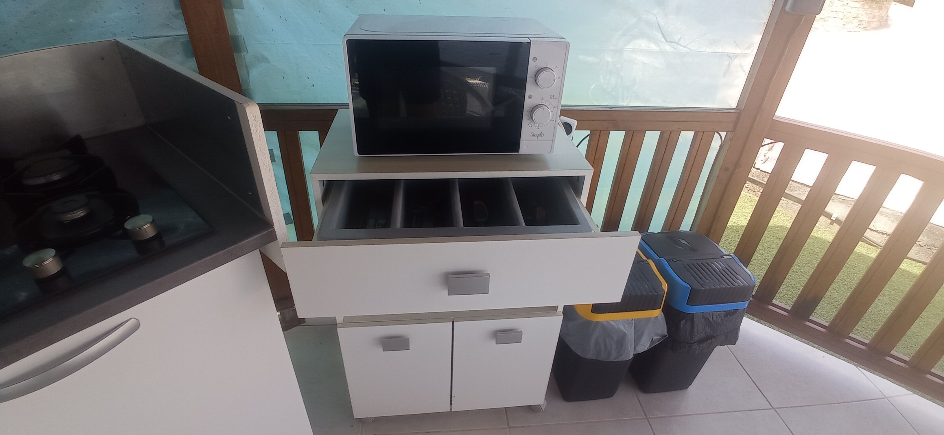 Une petite cuisine équipée d’un four à micro-ondes, d’une cuisinière à gaz et d’un petit réfrigérateur. On aperçoit deux poubelles et une rambarde en bois à l’arrière-plan.