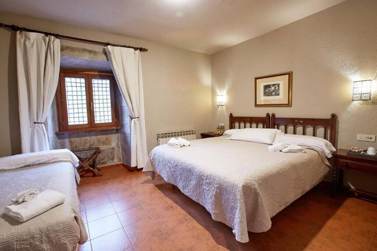 A bedroom with a large bed, a smaller bed, a window with curtains, a radiator, a nightstand, and a framed picture on the wall.