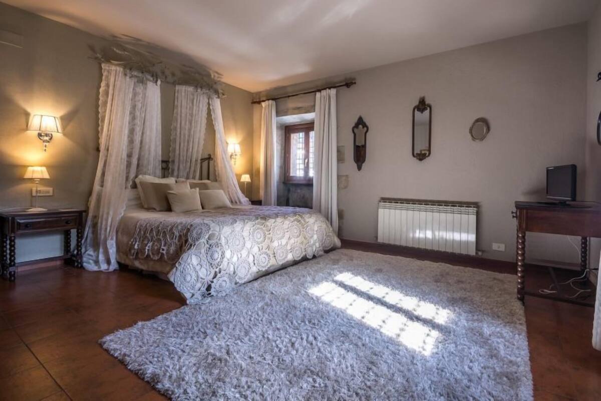 A bedroom with a four-poster bed, a large fluffy rug, a bedside table with a lamp, a radiator, and a TV on a wooden stand.